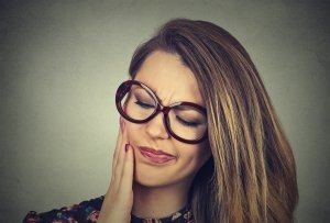 Closeup portrait young woman in glasses with sensitive toothache crown problem about to cry from pain touching outside mouth with hand isolated on gray background. Negative emotion face expression