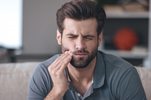 Frustrated young man touching his cheek and keeping eyes closed while sitting on the couch at home