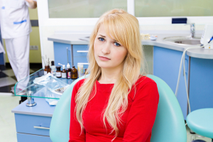 Closeup portrait sad, young girl, woman with painful tooth, ache in medical care office siting in chair, isolated dentist clinic office background. Human face expressions, emotions, feelings, reaction